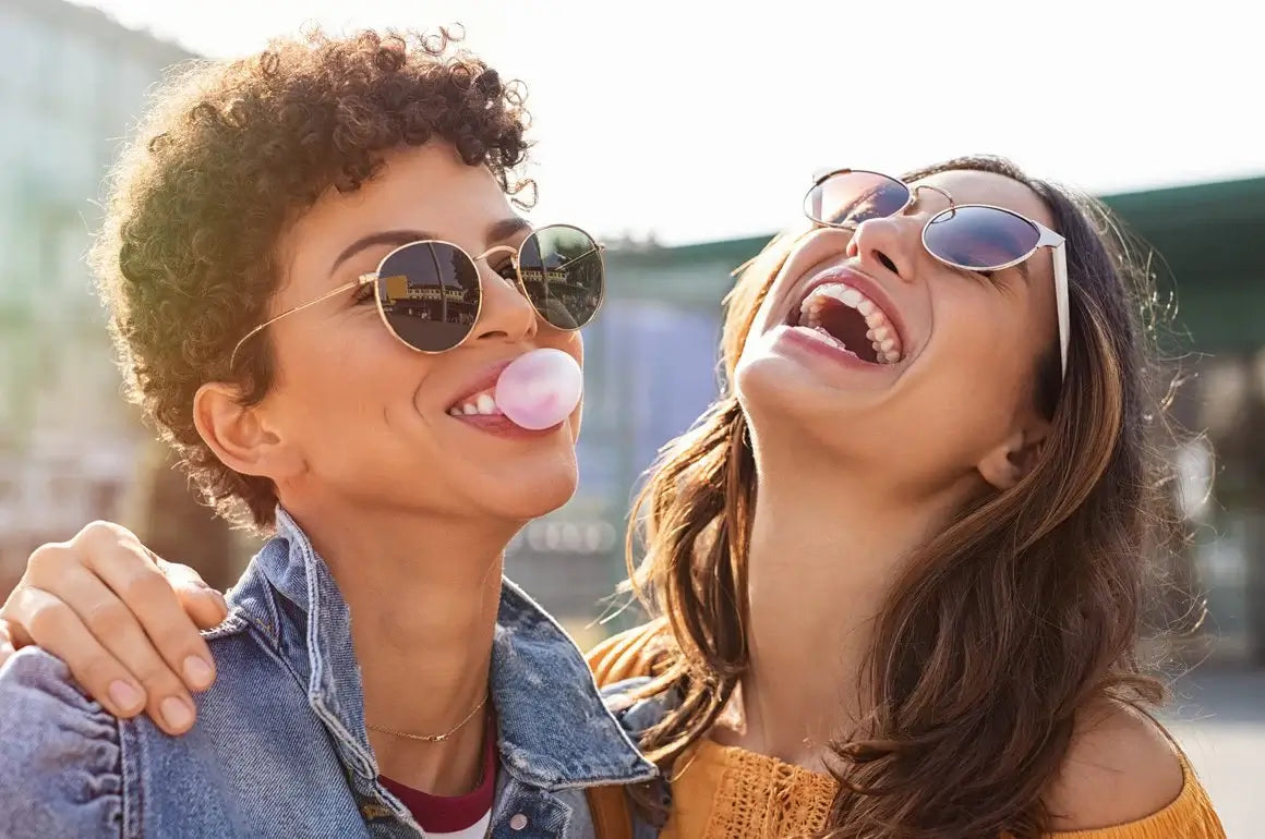 Two women laughing and smiling outdoors, one blowing a gum bubble.