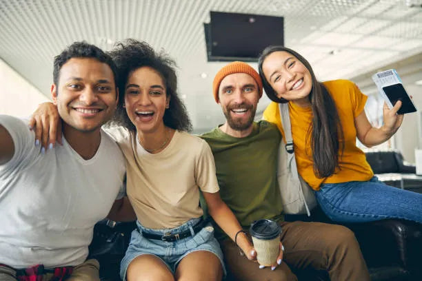 Group of friends smiling and posing for a selfie together at the airport, with luggage and departure signs in the background